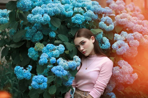 Image d'une jolie fille calme avec des cheveux bruns et un maquillage lumineux portant un t-shirt rose, debout parmi les fleurs bleues et regardant la caméra — Photo