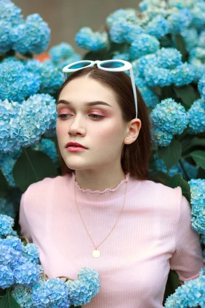 Retrato de una joven hermosa mujer atractiva con maquillaje tierno en camiseta rosa, con gafas de sol blancas en la cabeza de pie entre flores azules y mirando a la izquierda por la esquina —  Fotos de Stock