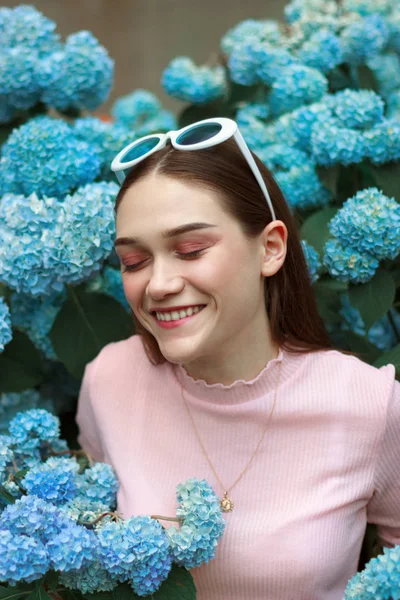 Feliz joven morena sonriente con maquillaje brillante y gafas de sol blancas en la cabeza, de pie entre flores azules con los ojos cerrados —  Fotos de Stock