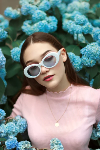 Elegante hermosa chica morena joven con labios rojos con camiseta de ping y gafas de sol blancas, de pie entre flores azules y mirando hacia abajo —  Fotos de Stock