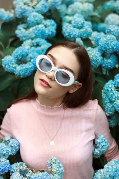 Chica morena en serio calma en camiseta rosa y gafas de sol blancas entre flores azules —  Fotos de Stock