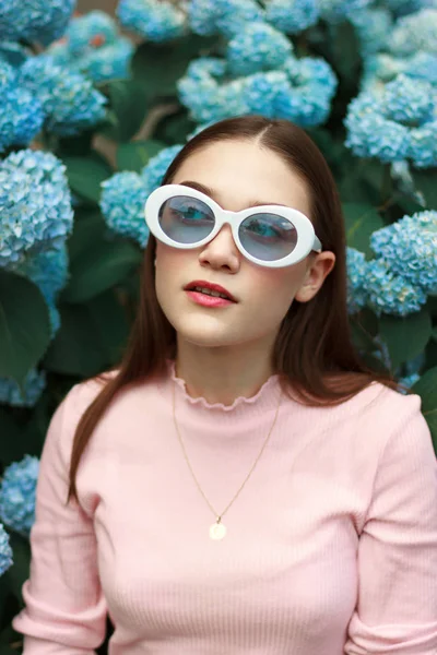 Chica morena en serio calma en camiseta rosa y gafas de sol blancas entre flores azules —  Fotos de Stock