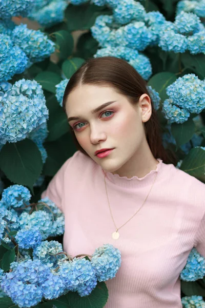 Retrato de belleza de una joven morena con maquillaje brillante, vestida con camiseta rosa de pie entre flores azules — Foto de Stock