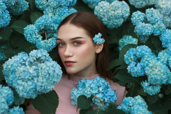 Gros plan visage de belle jeune femme aux lèvres rouges et aux yeux bleus et avec une fleur bleue derrière l'oreille, debout parmi les fleurs — Photo
