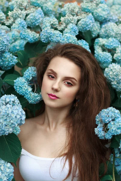 Sensuelle fille brune tendre aux cheveux lâches et aux lèvres rouges, vêtue de t-shirt blanc, debout parmi les fleurs bleues — Photo