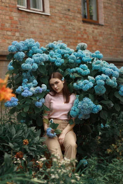 ピンクのTシャツとベージュのズボンを着た若い美しいブルネットの女の子は、カメラを見て青い花の間に座って — ストック写真