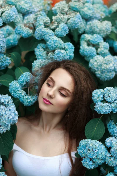 Czuły delikatny brunetka dziewczyna z luźnymi włosami i czerwonymi ustami, ubrana w białą koszulkę, stojąc wśród niebieskich kwiatów — Zdjęcie stockowe