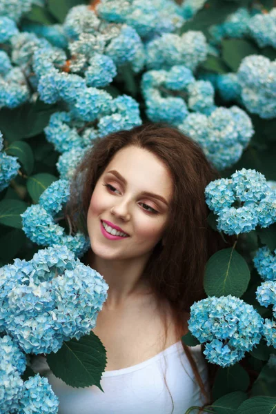 Joyeux sourire fille brune avec des lèvres rouges en t-shirt blanc debout parmi les fleurs bleues — Photo