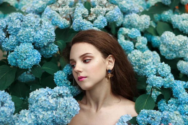 Magnifique jeune fille brune à la mode avec une peau naturelle saine et des lèvres rouges, avec des épaules nues debout parmi les fleurs bleues — Photo