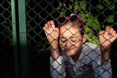 Başını pembe güneş gözlüğü ile sevimli güzel kız kapalı gözleri ile ızgara arkasında durmak, ellerini tutarak, gece