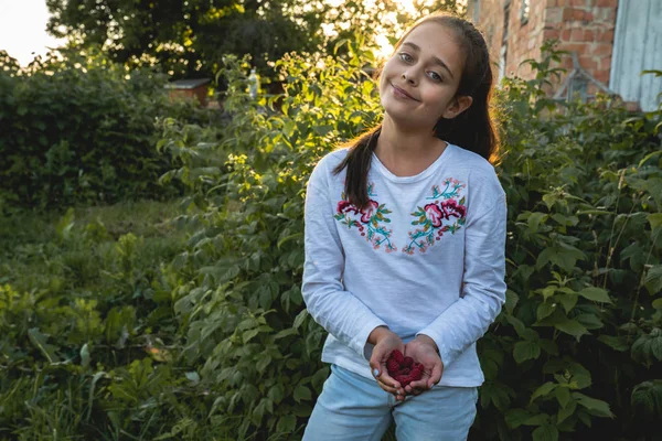 Feliz chica morena sonriente mostrando frambuesa en las palmas de las manos que se reunieron en el patio trasero, vacaciones de verano — Foto de Stock