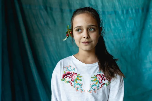 Jeune fille mignonne sérieusement avec des cheveux bruns en t-shirt blanc avec broderie nationale avec une branche de viorne derrière l'oreille, regardant loin isolé sur fond bleu — Photo