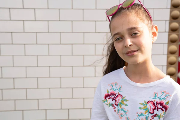 Petite fille brune joyeuse avec une peau saine regardant calmement la caméra avec des lunettes de soleil roses sur la tête, isolée sur un mur de briques blanches — Photo