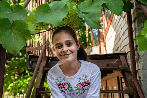 Joyeux sourire fille brune en t-shirt blanc assis sur un escalier en métal et regardant la caméra, regardant hors des branches de vigne — Photo