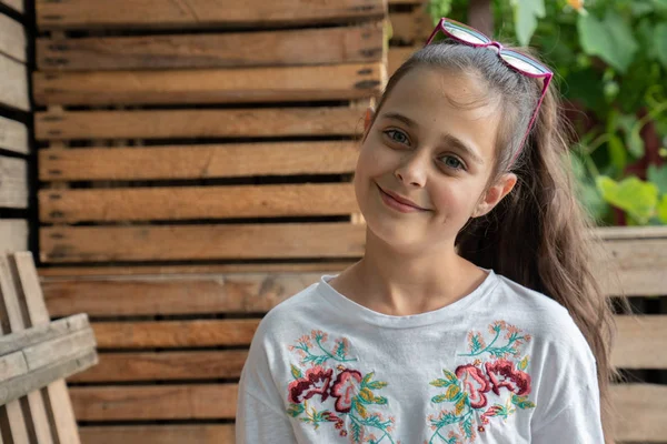 Jolie petite fille heureuse en t-shirt blanc avec des lunettes de soleil roses sur la tête en regardant la caméra, des boîtes en bois sur le fond — Photo