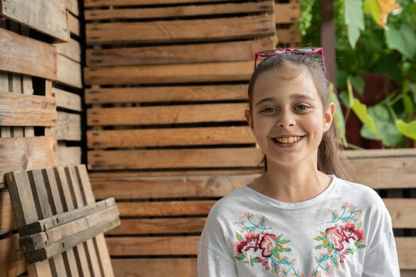 Jolie petite fille heureuse en t-shirt blanc avec des lunettes de soleil roses sur la tête en regardant la caméra, des boîtes en bois sur le fond — Photo