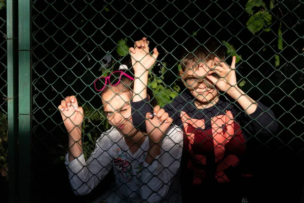 Due bambini in abiti casual in piedi dietro la griglia, facendo facce divertenti con un'espressione spaventosa, di notte — Foto Stock