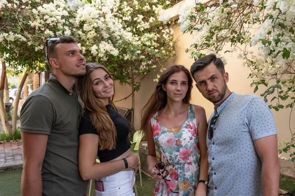 Young friends, two brunette girls and to attractive boys in polo t-shirts standing against wall with beautiful white flowers — Stock Photo, Image