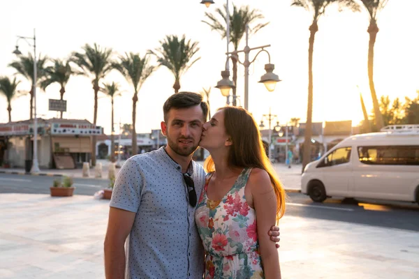 Lovely couple, brunetet girl in dress with flowers print kissing her boyfriend in blue shirt while standing on the embankment street with palms, sunset on the background — Stock Photo, Image