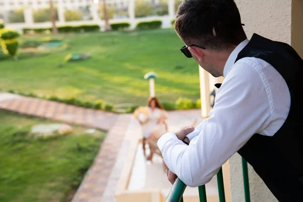 Young man in white shirt and sunglasses leaned on the balcony railing, on blurred background beautiful girl in white dress sitting on chair of the summer terrace