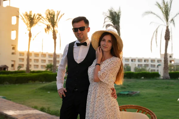 Feliz joven pareja, hombre de camisa blanca y gafas de sol abrazando a su novia en sombrero y vestido delicado, ambos mirando a la cámara, puesta de sol en el fondo — Foto de Stock