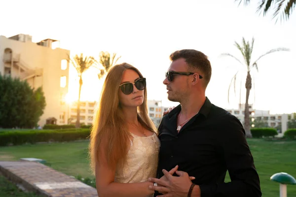 Half length portrait of young serious couple, man in black shirt hugging his blonde girlfriend in delicate dress, both in sunglasses, sunset on the background — Stock Photo, Image