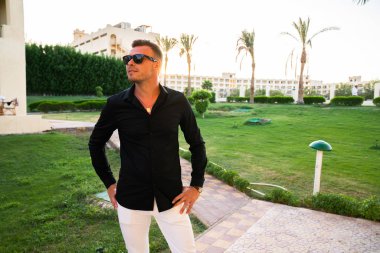 Güneş gözlüğü giyen genç adam kemerini el ele tutuşarak ve uzağa bakarak, arka planda gün batımı