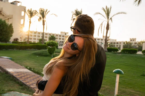 Joven hombre de camisa negra disfrutando de la puesta del sol y abrazando a su novia que mirando a la cámara sobre su hombro — Foto de Stock