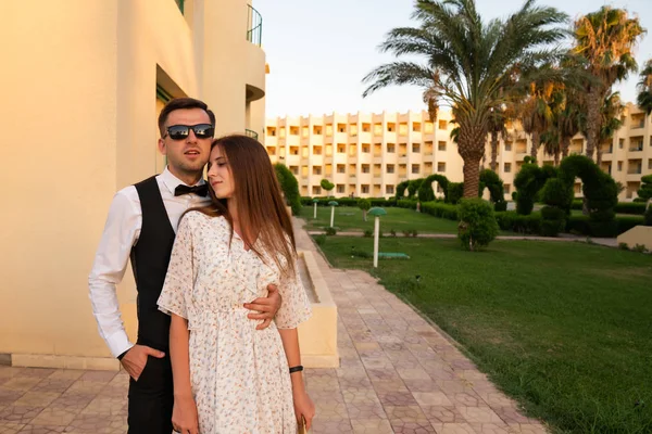 Joven hombre atractivo en camisa blanca y gafas de sol abrazando a su hermosa novia morena en vestido delicado, área de hotel bien cuidada en el fondo — Foto de Stock