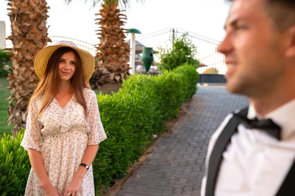 Giovane uomo sfocato in camicia bianca, la sua ragazza in abito delicato e cappello in piedi accanto e lo guarda — Foto Stock