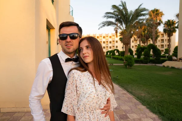 Jeune homme attrayant en chemise blanche et lunettes de soleil embrassant sa belle petite amie brune en robe délicate, zone de l'hôtel bien entretenu sur le fond — Photo