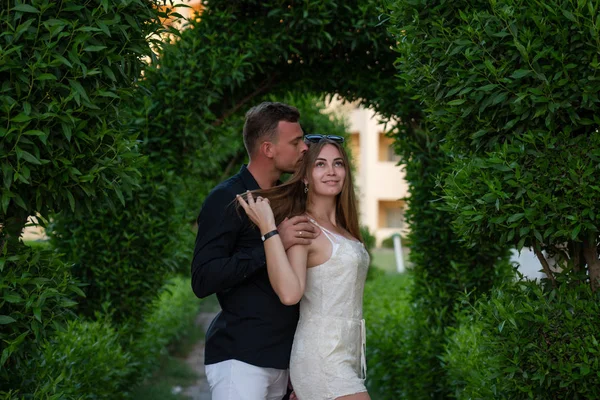 Handsome young man in black shirt hugs his gierlfriend in white dress, green leaves wall on the background — Stock Photo, Image