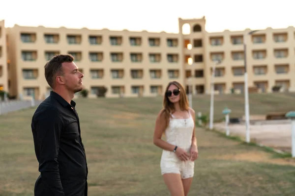 Hamdsome young man in black shirt looking straight, her girlfriend in sunglasses and white dress standing beside and looking at him