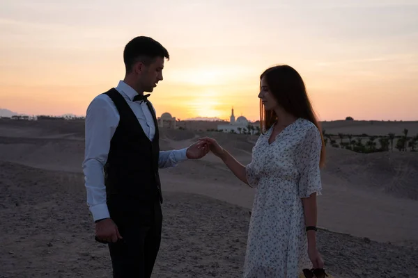 Romantic couple in elegant clothes standing among the desert opposite each other and holding hand in hand, amazing sunset on the background — Stock Photo, Image