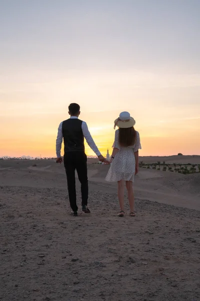 Beautiful couple in elegant clothes holding hand in hand, walking among the desert, enjoying amazing sunset together — Stock Photo, Image