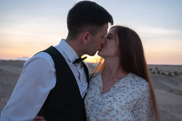 Beautiful young couple in elegant clothes kissing at sunset among the desert — Stock Photo, Image