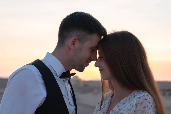 Romantic couple, brunette girl in delicate dress and handsome man in white shirt pressed their heads to each other at sunset among the desert — Stock Photo, Image