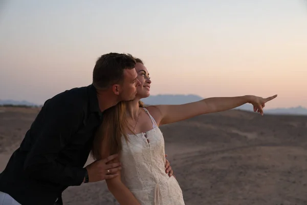 Handsome young man hugging his girlfriend in white dress, she pointing and looking into the distance, couple standing among the desert — Stock Photo, Image