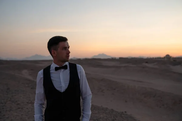 Joven serio con camisa blanca de pie entre el desierto con las manos en los bolsillos y mirando hacia otro lado, puesta de sol en el fondo — Foto de Stock