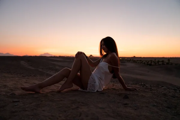 Misteriosa ragazza bruna in abito bianco seduta sulla sabbia tra il deserto, il tramonto e il cielo colorato sullo sfondo — Foto Stock