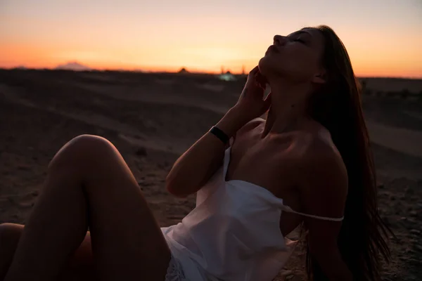 Fille brune ludique en robe blanche avec des épaules nues assis sur le sable au milieu du désert et touchant son visage avec la main, ciel coloré sur le fond — Photo