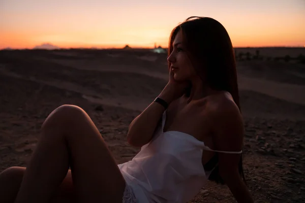 Menina morena brincalhão em vestido branco com ombros nus sentados na areia entre o deserto e tocando seu rosto com a mão, céu colorido no fundo — Fotografia de Stock