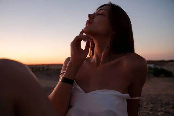 Close up retrato de bela jovem com cabelos morena e ombros nus que sentado na areia entre o deserto tocando seu rosto com a mão, pôr do sol no fundo — Fotografia de Stock