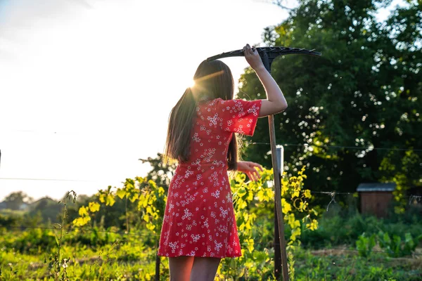 Девочка-подросток с брюнеткой в красном платье, стоящей спиной к камере, опираясь на грабли, отдыхая во время работы в винограднике, закат на заднем плане, люди и садовая концепция — стоковое фото
