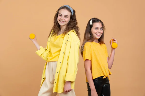 Två små lekfulla barn i gula skjortor, systrar eller tonåriga biracial flickor med brunt lockigt hår spelar eller tränar, lyfta hantlar — Stockfoto