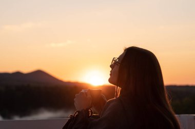 Kapalı gözlü bir kız gün batımında kahve içip manzarayı seyrediyor.