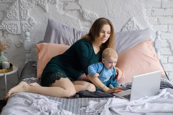美しい若い母親と幼い息子はベッドに座るし ノート パソコンの本で興味を持って傍観 — ストック写真