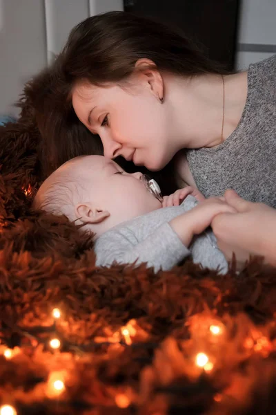 一位年轻的母亲看着她的孩子 她睡在蓬松的棕色毯子上 周围都铺着灯光 — 图库照片