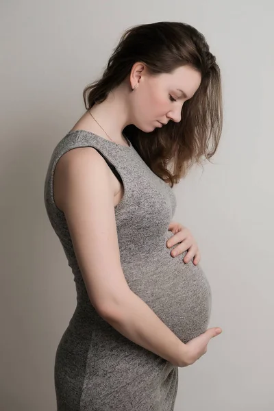 グレーのドレス ホワイト バック グラウンドに立って 彼の腹を見て 若い妊娠中の女の子 — ストック写真