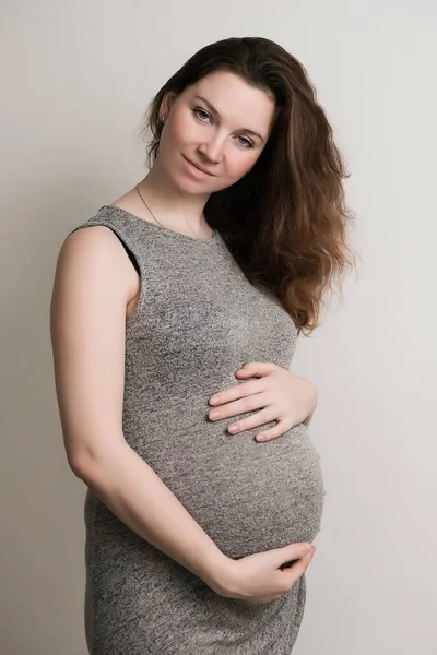 一个年轻的怀孕女孩在灰色的礼服站在白色的背景 它拥抱你的肚子 — 图库照片